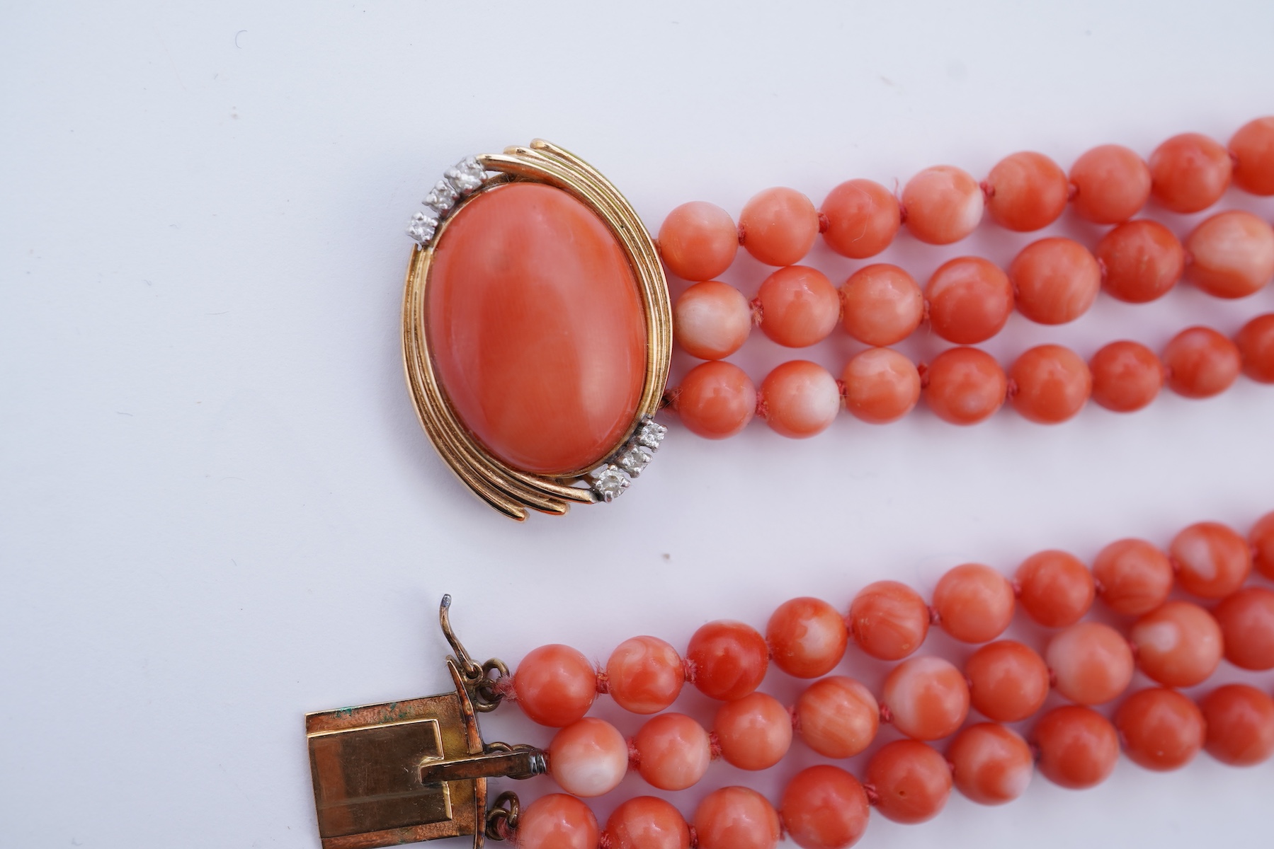 A coral necklace, composed of three strands of coral beads measuring approximately 5.1-6.6mm diameter, to a gold clasp set with a coral cabochon and brilliant-cut diamonds, mounted in gold, length 45.5cm, stamped 14K, ca